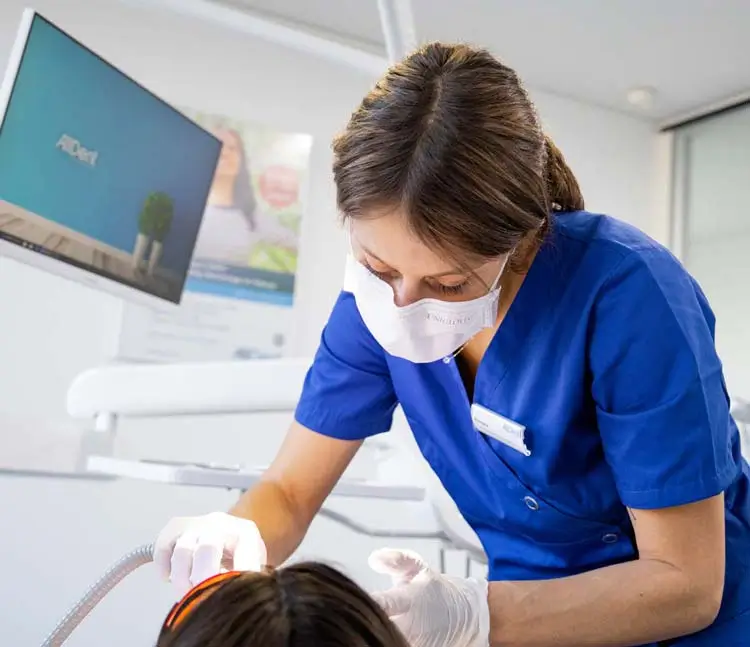 Zahnärztin in blauer Arbeitskleidung und Maske behandelt eine Patientin im Behandlungsstuhl, im Hintergrund moderner Bildschirm