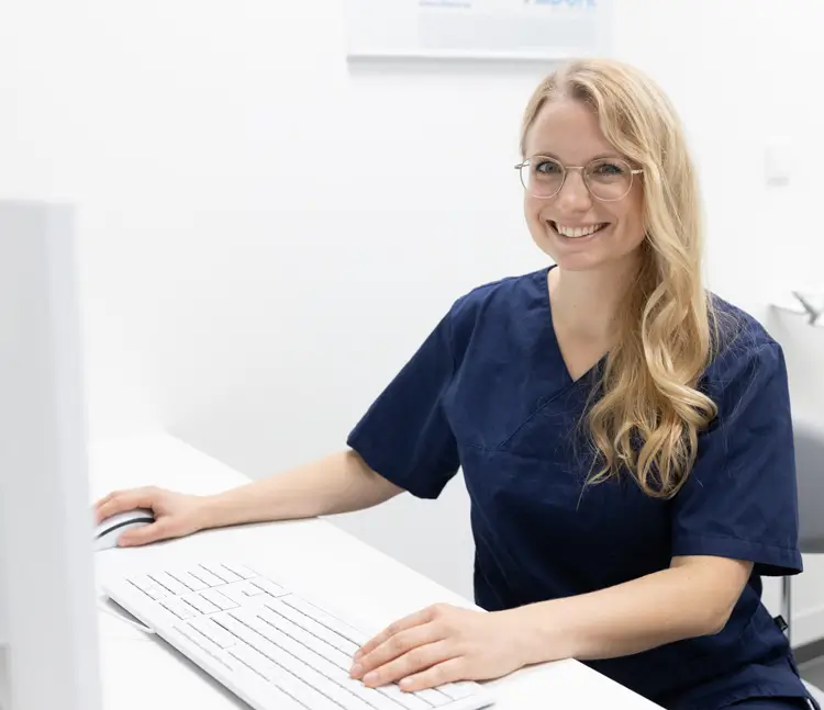 Eine Frau in blauer Arbeitskleidung sitzt an einem Schreibtisch vor einem Computer, hält eine Hand auf der Maus und lächelt in die Kamera.