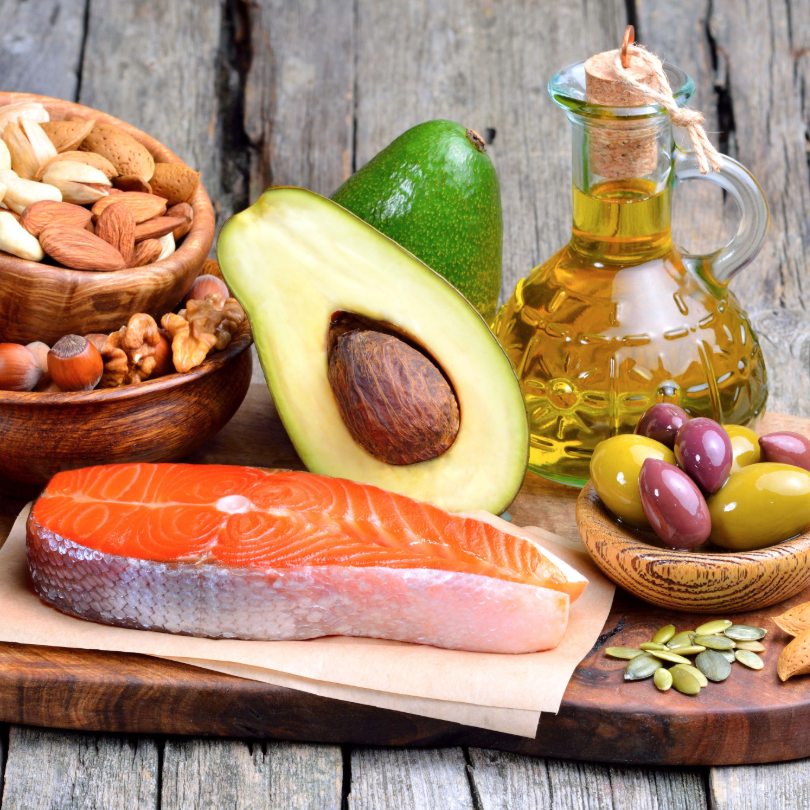 Halbierte Avocado, rohes Lachsfilet, Oliven&ouml;l in Glasflasche, verschiedene N&uuml;sse und gr&uuml;ne sowie violette Oliven auf Holzbrett