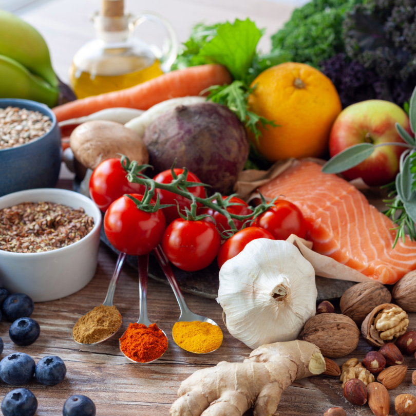 Verschiedene frische Lebensmittel auf Holztisch, darunter Kirschtomaten, Knoblauch, Ingwer, Karotte, Lachsfilet, Apfel, Orange, N&uuml;sse, Haferflocken, Gew&uuml;rze und Oliven&ouml;l