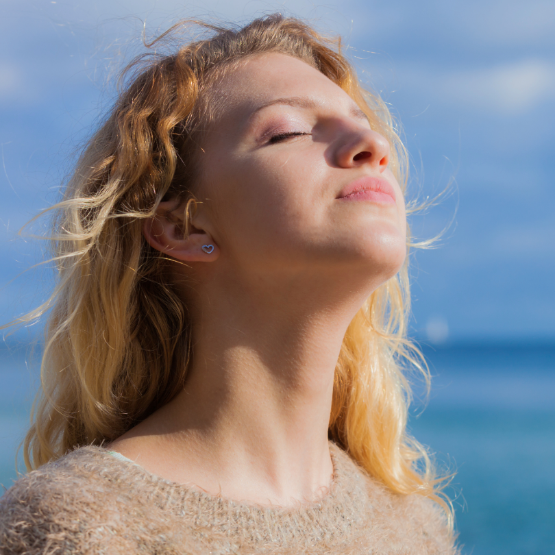 Frau mit langen, lockigen blonden Haaren tr&auml;gt einen beigen Pullover, Blick nach oben zum blauen Himmel am Meer