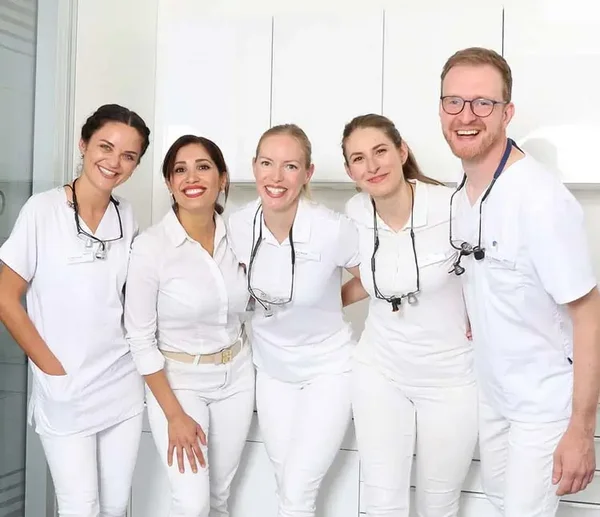 Team aus Zahn&auml;rzten steht nebeneinander im Behandlungszimmer und l&auml;chelt in die Kamera
