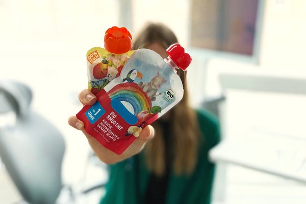 Person hält zwei bunte Quetschbeutel mit Bio-Smoothies in die Kamera, im Hintergrund unscharf eine Zahnarztpraxis