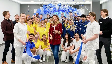 Gruppe von Menschen in gelben, blauen und wei&szlig;en Arbeitskleidungen bei einer Er&ouml;ffnungsfeier mit blauen Luftballons und Konfetti.