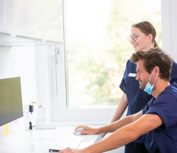 Ein Mann und eine Frau in blauer Arbeitskleidung stehen an einem Schreibtisch und schauen auf einen Computerbildschirm