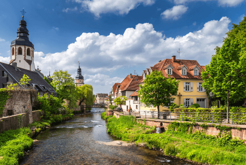 Blick auf die Pfinz, links des Flusses eine Kirche, rechts ein großes Gebäude